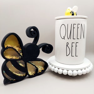 May include: Black and gold glitter bee cutout with a white ceramic jar with the text "Queen Bee" and a small yellow and black bee figurine on top.