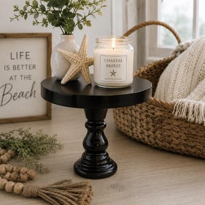 May include: A black pedestal stand holding a glass jar candle labeled Coastal Breeze with a starfish icon. Next to the candle is a beige starfish in a white vase with greenery. A wooden bead garland with a tassel lies in the foreground.