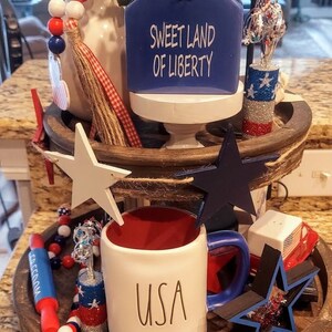 May include: A two-tiered wooden tray decorated with patriotic items including a blue house-shaped sign with the text SWEET LAND OF LIBERTY, a white mug with USA printed on it, wooden stars in white and blue, red white and blue beads, and small glittery accents.