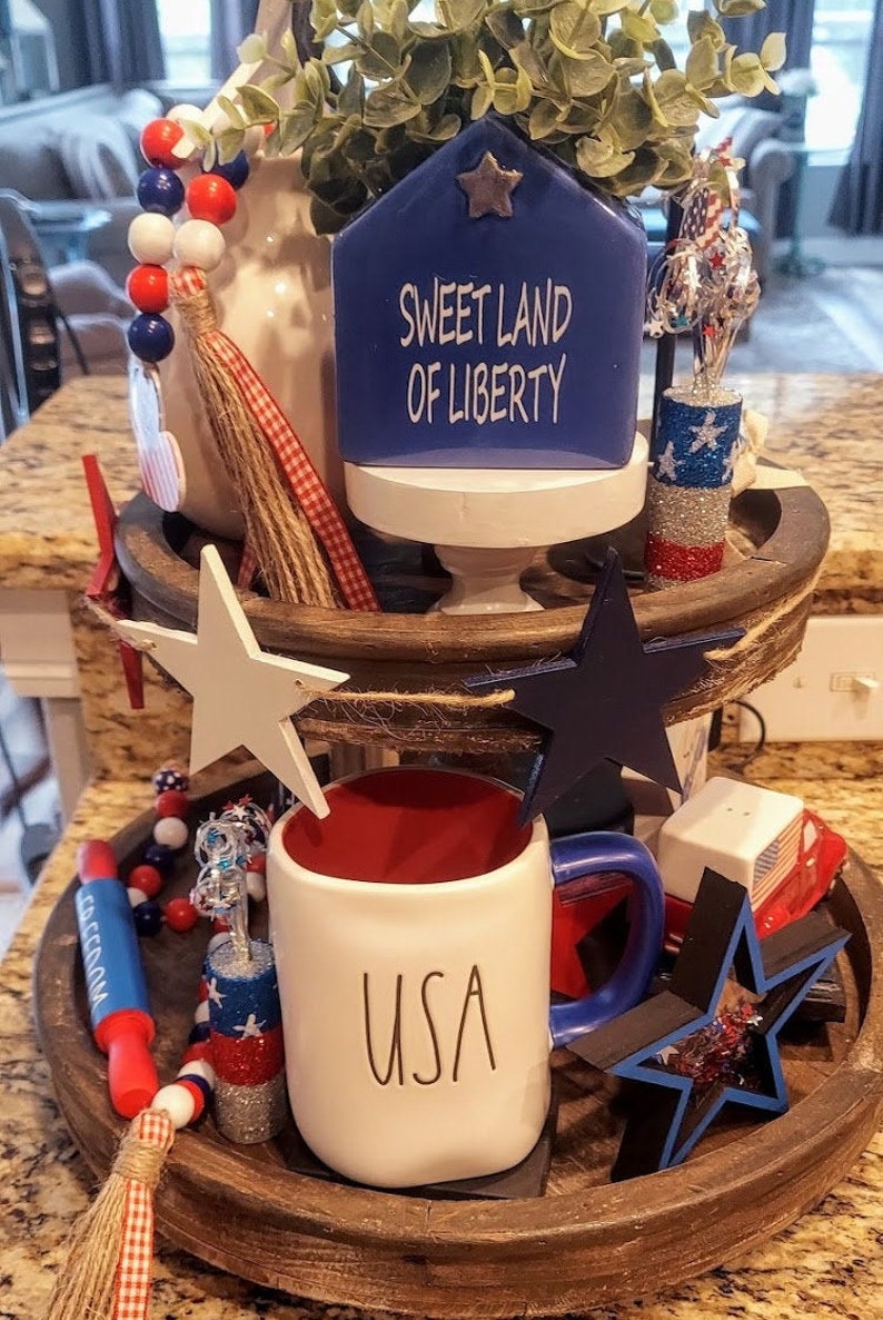 May include: A two-tiered wooden tray decorated with patriotic items including a blue house-shaped sign with the text SWEET LAND OF LIBERTY, a white mug with USA printed on it, wooden stars in white and blue, red white and blue beads, and small glittery accents.