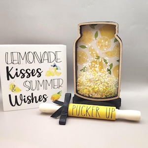 May include: A decorative wooden sign with the text "Lemonade Kisses & Summer Wishes" next to a jar-shaped decoration filled with lemon slices and glitter. A yellow rolling pin with the words "Pucker Up" is tied with a black ribbon.