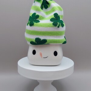 May include: A white ceramic mug with a simple face featuring two black eyes and a small orange nose, topped with a knit hat featuring green and white horizontal stripes, dark green shamrock patterns, and a small green satin bow on top.