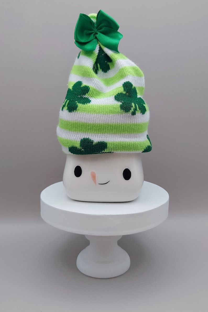May include: A white ceramic mug with a simple face featuring two black eyes and a small orange nose, topped with a knit hat featuring green and white horizontal stripes, dark green shamrock patterns, and a small green satin bow on top.