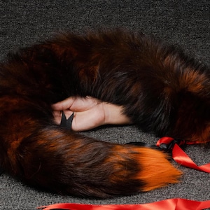 May include: A long, fluffy, brown and orange faux fur tail with a black ribbon tied around it. The tail is curled up and held in a hand.
