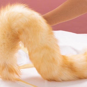 Puede incluir: Una cola de piel sintética esponjosa de color marrón claro con una punta larga y rizada. La cola está sobre una superficie blanca.