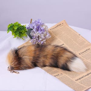 May include: A fluffy brown, black, and white faux fur tail keyring with a gold metal ring. The tail is on a white surface with a newspaper underneath.