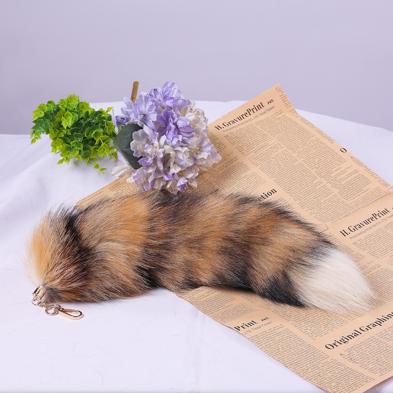 May include: A fluffy brown, black, and white faux fur tail keyring with a gold metal ring. The tail is on a white surface with a newspaper underneath.