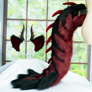 May include: A costume set featuring a headband with black and red pointed ears and curved horns, paired with a long, plush tail. The tail is covered in black and deep red faux fur with a row of red spikes running along the top edge.