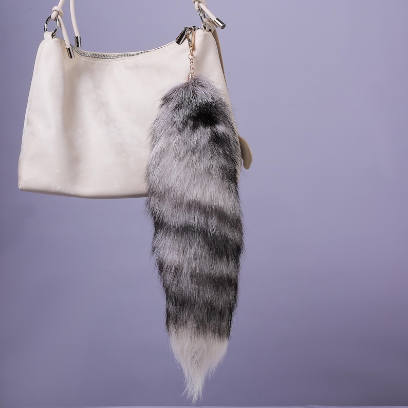 May include: A white faux leather handbag with a silver chain and a fluffy grey and white fox tail keychain.