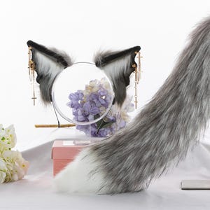 May include: A gray and white faux fur tail with a white tip, a headband with gray and white faux fur cat ears, and gold dangling charms. The headband and tail are displayed on a white surface with a pink box and white flowers.