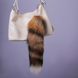 May include: A brown and white faux fur tail key ring hanging from a white handbag. The key ring is attached to a gold metal ring.