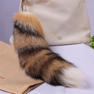 May include: A fluffy brown, black and white faux fur tail keychain with a gold chain. The tail is attached to a light brown leather handbag with the text "CHLO and CHLO" on the front.