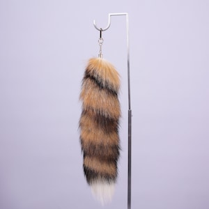 May include: A fluffy brown and black faux fur tail keyring with a white tip. The tail is attached to a gold-coloured metal ring.