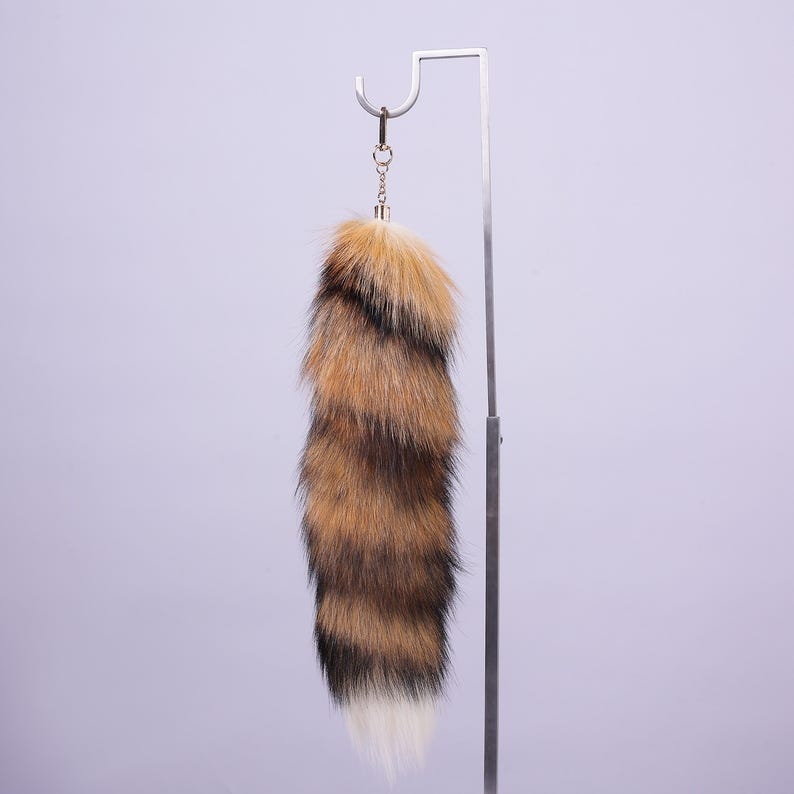May include: A fluffy brown and black faux fur tail keyring with a white tip. The tail is attached to a gold-coloured metal ring.