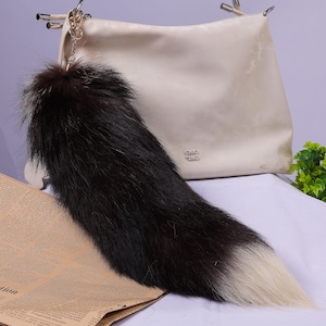 May include: A black and white faux fur tail keychain attached to a white handbag. The keychain is made of soft, fluffy fur and has a long, flowing tail.