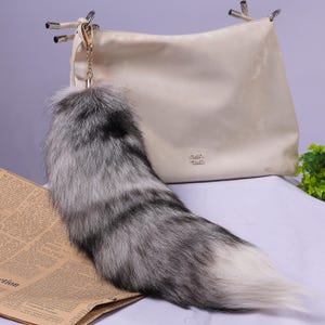 May include: A fluffy grey and white fox tail keyring attached to a gold chain. The keyring is hanging from a cream-coloured handbag.