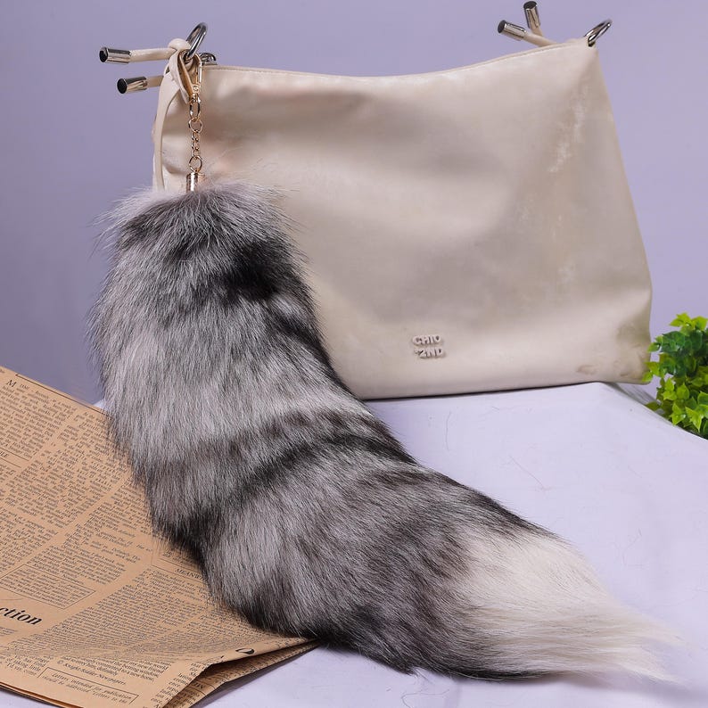 May include: A fluffy grey and white fox tail keyring attached to a gold chain. The keyring is hanging from a cream-coloured handbag.
