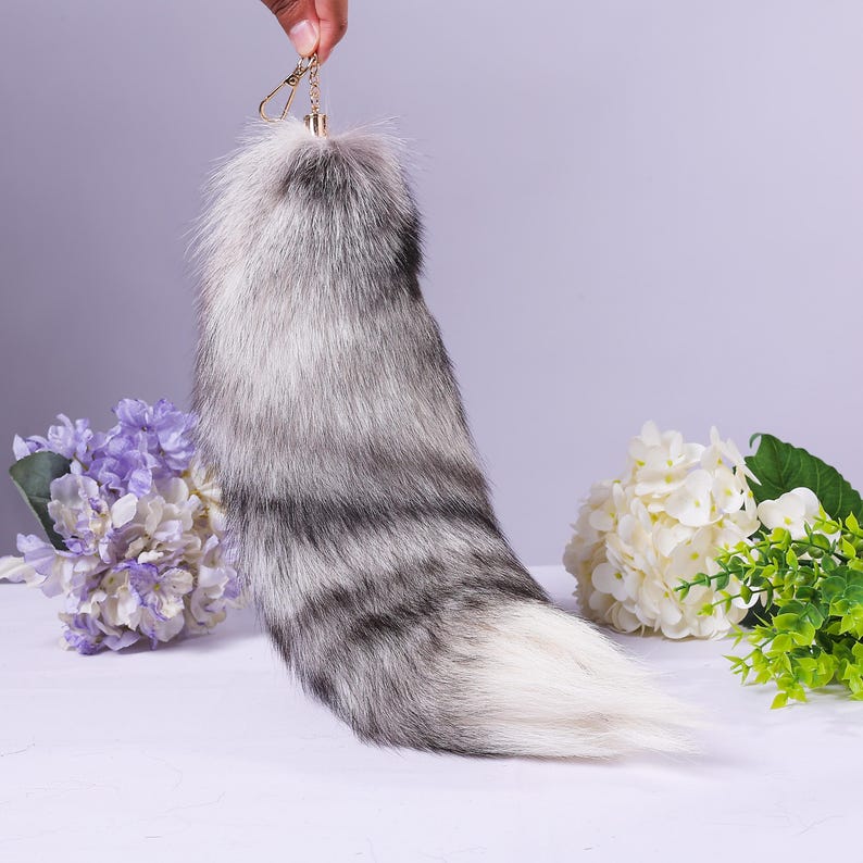 May include: A fluffy white and grey faux fur tail keyring with a gold metal ring. The tail is long and bushy, and it is perfect for adding a touch of whimsy to any bag or keyring.