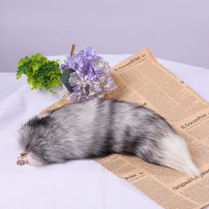 May include: A fluffy grey and white faux fur tail with a gold chain attached. The tail is lying on a newspaper with a bouquet of purple flowers in the background.