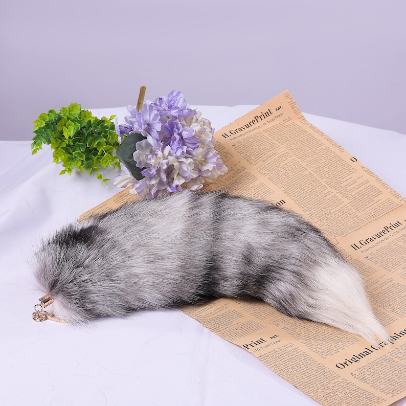 May include: A fluffy grey and white faux fur tail with a gold chain attached. The tail is lying on a newspaper with a bouquet of purple flowers in the background.