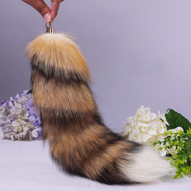 May include: A fluffy, multi-coloured fox tail keyring with a gold metal ring. The tail is made of faux fur and has a white tip. The tail is curled up and is sitting on a white surface.