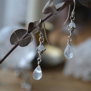 May include: A pair of silver dangle earrings with teardrop-shaped white moonstone beads. The earrings are hanging from a branch.