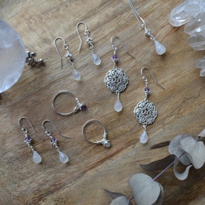 May include: A collection of silver earrings with moonstone drops and amethyst accents. The earrings are displayed on a wooden surface with a clear quartz crystal and a eucalyptus branch.