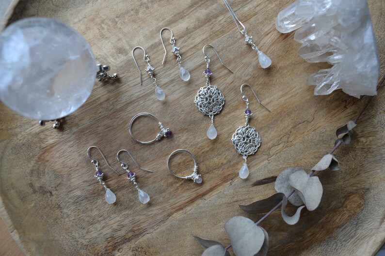 May include: A collection of silver earrings with moonstone drops and amethyst accents. The earrings are displayed on a wooden surface with a clear quartz crystal and a eucalyptus branch.