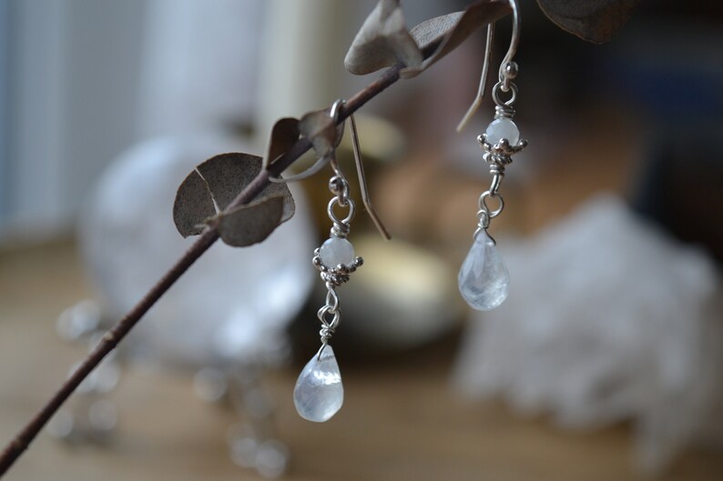 May include: A pair of silver dangle earrings with teardrop-shaped white moonstone beads. The earrings are hanging from a branch.