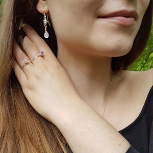 May include: A woman wearing a black shirt and a silver necklace with two silver rings on her fingers. One ring has a small purple stone and the other has a white stone. She is also wearing silver earrings with a teardrop-shaped white stone.