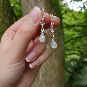 May include: Silver dangle earrings with a teardrop-shaped moonstone and a small amethyst gemstone.
