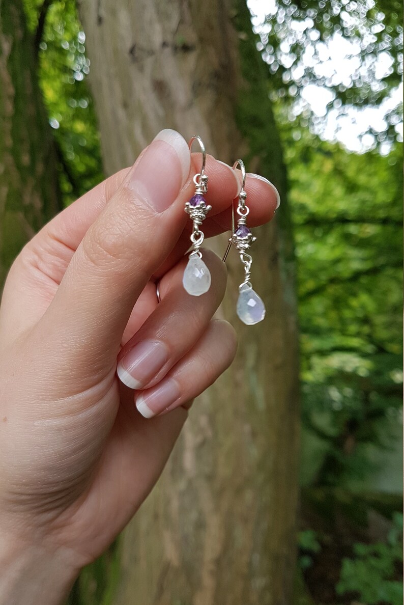May include: Silver dangle earrings with a teardrop-shaped moonstone and a small amethyst gemstone.