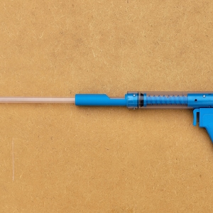 May include: A blue and clear plastic tool with a long, clear barrel and a blue handle. The tool has a spring inside the barrel and a small blue nozzle at the end.