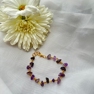 May include: A gold-colored bracelet with small, irregular purple stones and gold beads. The bracelet is displayed on a white surface next to a white chrysanthemum flower.