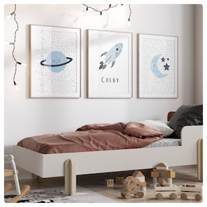 May include: Three framed art prints with a space theme above a white bed. The prints depict a planet, a rocket with the name "Colby", and a moon with stars. The bed has a brown duvet and a grey pillow. The room is decorated with fairy lights.