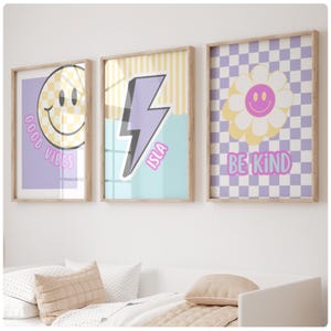 Könnte beinhalten: Drei gerahmte Wandkunstdrucke mit hellem Holzfinish. Die Drucke zeigen ein Smiley-Gesicht, einen Blitz mit dem Namen "ISLA" und eine Gänseblümchen mit einem Smiley-Gesicht in der Mitte. Jeder Druck hat ein einzigartiges Hintergrunddesign und positive Botschaften.