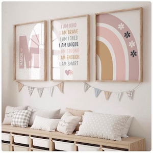 May include: Three framed prints with a pink and brown color scheme. The first print features the letter "M" with the name "MARCIE" written vertically. The second print has the text "I AM KIND, I AM BRAVE, I AM LOVED, I AM UNIQUE, I AM STRONG, I AM ENOUGH, I AM SMART". The third print features a rainbow with pink, brown, and white stripes and black flowers.