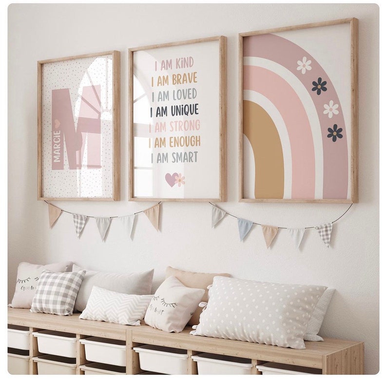 May include: Three framed prints with a pink and brown color scheme. The first print features the letter "M" with the name "MARCIE" written vertically. The second print has the text "I AM KIND, I AM BRAVE, I AM LOVED, I AM UNIQUE, I AM STRONG, I AM ENOUGH, I AM SMART". The third print features a rainbow with pink, brown, and white stripes and black flowers.