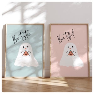 May include: Two framed prints with a ghost holding a pumpkin. The print on the left says "Boo-tastie" in black script on a light blue background. The print on the right says "Boo-tiful" in black script on a light pink background.