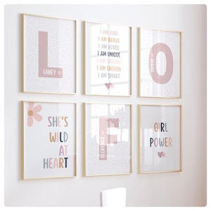 May include: Six framed prints with gold frames and white backgrounds. The prints feature pink and white polka dots, pink and white flowers, and pink letters. The prints have the following text: "L Laney", "I am kind I am brave I am loved I am unique I am strong I am enough I am smart", "She's wild at heart", "Florence", "O Olivia", and "Girl Power".