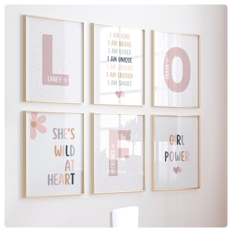 May include: Six framed prints with gold frames and white backgrounds. The prints feature pink and white polka dots, pink and white flowers, and pink letters. The prints have the following text: "L Laney", "I am kind I am brave I am loved I am unique I am strong I am enough I am smart", "She's wild at heart", "Florence", "O Olivia", and "Girl Power".