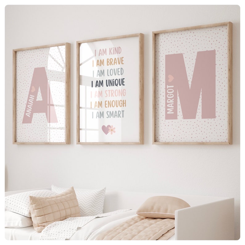 May include: Three framed prints with pink and white polka dot backgrounds. The left print features the letter A with the name Anaiah written vertically. The middle print features the words "I am kind, I am brave, I am loved, I am unique, I am strong, I am enough, I am smart" with a pink heart and a white flower. The right print features the letter M with the name Margot written vertically.