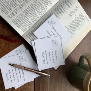 Puede incluir: Primer plano de una mesa de madera con una taza verde, un bolígrafo y varias tarjetas blancas con citas inspiradoras y versículos bíblicos. Las tarjetas presentan diseños florales y el texto "Ofrece el sacrificio de alabanza" y "Es mejor confiar en el Señor que confiar en el hombre. Salmo 118:8".