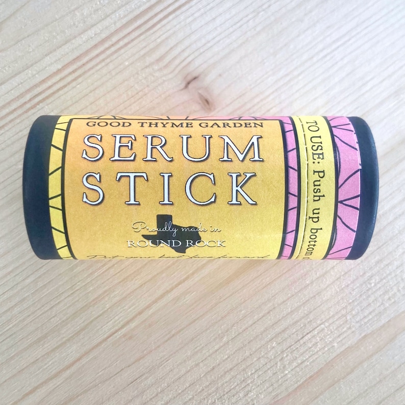 May include: A cylindrical serum stick with a yellow label. The label features the text "SERUM STICK" in large white letters, along with "GOOD THYME GARDEN" and "Proudly made in ROUND ROCK". The stick is black with pink floral accents.