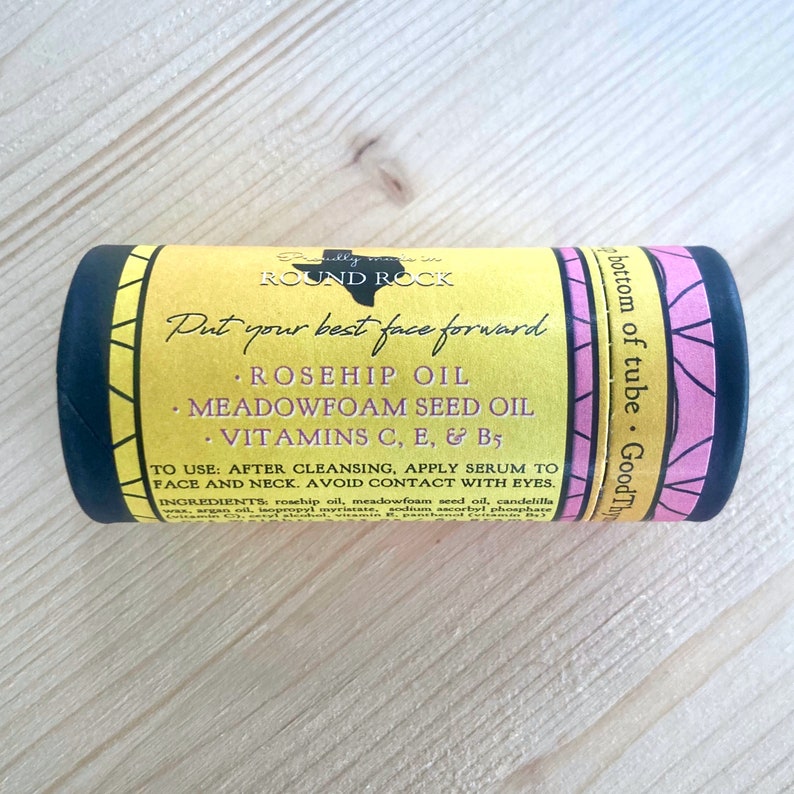 May include: A cylindrical skincare product tube with a yellow label featuring the text "ROUND ROCK" and "Put your best face forward." The label lists ingredients like rosehip oil and vitamins. The tube has black and pink accents.