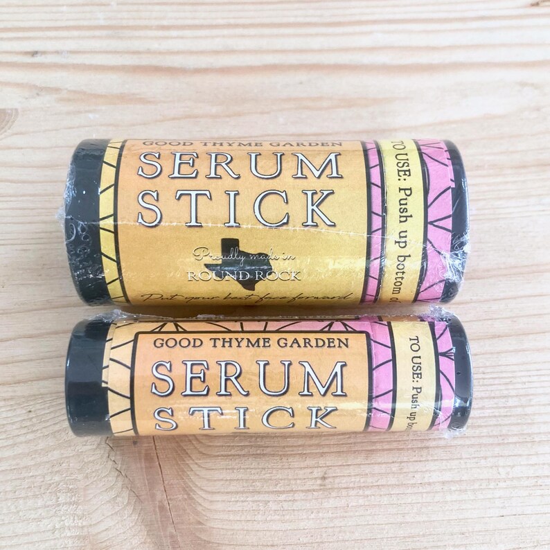 May include: Two cylindrical serum sticks in clear wrapping. The labels are tan with black text: "GOOD THYME GARDEN SERUM STICK". The ends are black. The text "TO USE: Push up bottom" is also visible on the label.