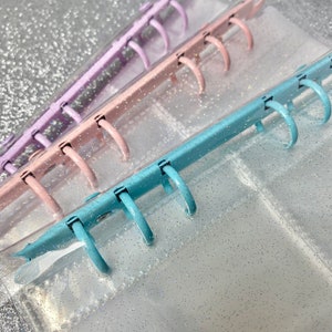 May include: Three clear plastic binders with glitter embedded in the plastic. The binders are stacked on top of each other. The top binder is light blue, the middle binder is pink, and the bottom binder is purple. Each binder has six metal rings.