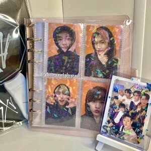 K&ouml;nnte beinhalten: Ein durchsichtiger Plastikordner mit glitzernder Oberfl&auml;che enth&auml;lt Foto-Karten einer K-Pop-Boyband. Der Ordner hat einen goldenen Ringmechanismus und eine durchsichtige Plastik-Tasche auf der Vorderseite. Die Foto-Karten zeigen Bilder der Bandmitglieder mit einem Konfetti-Hintergrund. Der Ordner steht auf einem Tisch neben einem kleinen wei&szlig;en Staffelei, auf dem eine Foto-Karte der Band steht.