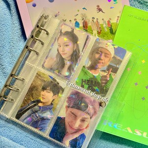 K&ouml;nnte beinhalten: Ein durchsichtiger Plastikordner mit glitzernder Oberfl&auml;che enth&auml;lt Foto-Karten von verschiedenen K-Pop-Idolen. Der Ordner hat einen silbernen Ringmechanismus und ist mit durchsichtigen Plastikh&uuml;llen gef&uuml;llt. Die Foto-Karten zeigen Bilder der Idolen in verschiedenen Posen und Umgebungen. Der Ordner ist ge&ouml;ffnet, um die Foto-Karten zu zeigen, die in Reihen angeordnet sind. Der Ordner liegt auf einer gr&uuml;nen Oberfl&auml;che mit wei&szlig;em Hintergrund.