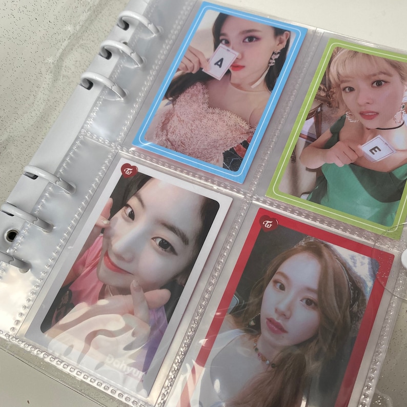 May include: A clear plastic binder with a collection of photo cards featuring a K-pop girl group. The cards are in clear plastic sleeves and feature different members of the group. The cards have a pink and white heart logo with the text "Twice" on them. The cards feature the letters "A" and "E" on them.